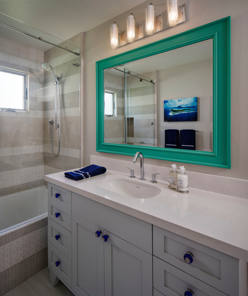 Maritim inredning av ett litet badrum med dusch, med skåp i shakerstil, blå skåp, ett badkar i en alkov, en dusch/badkar-kombination, beige kakel, keramikplattor, beige väggar, ett undermonterad handfat och bänkskiva i akrylsten
