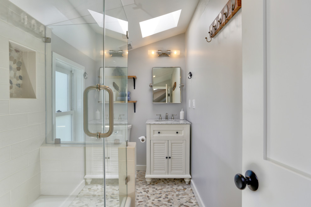 Пример оригинального дизайна: ванная комната в морском стиле с фасадами с декоративным кантом, белыми фасадами, ванной в нише, душем над ванной, бежевой плиткой, серыми стенами, полом из галечной плитки, столешницей из гранита, разноцветным полом и душем с распашными дверями