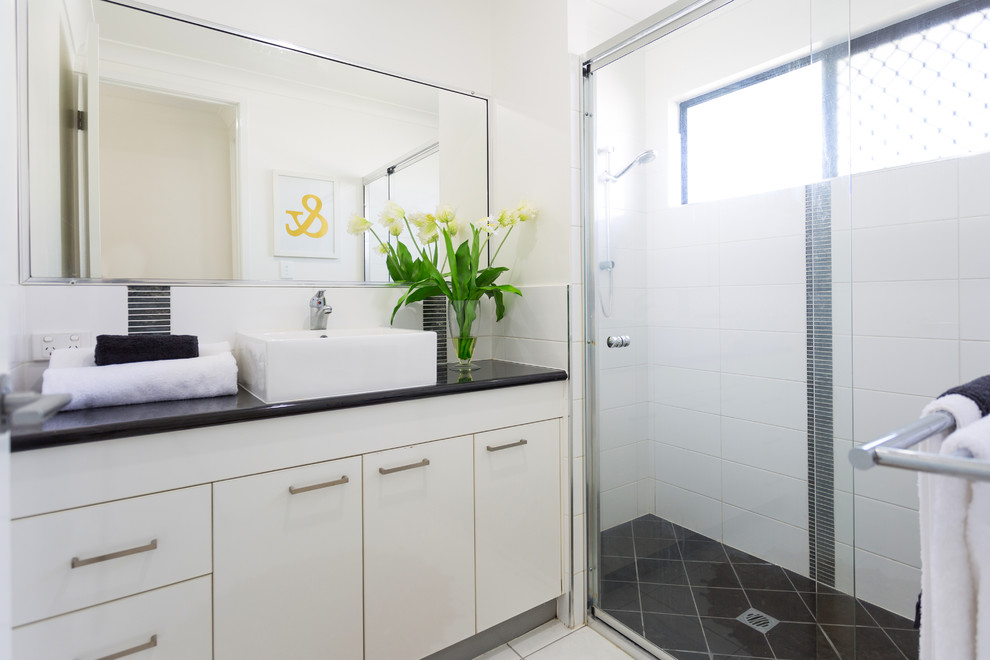 Cette photo montre une salle de bain bord de mer avec un placard à porte plane, des portes de placard beiges, un carrelage blanc, un mur blanc, une vasque, un sol beige, une cabine de douche à porte coulissante et un plan de toilette noir.
