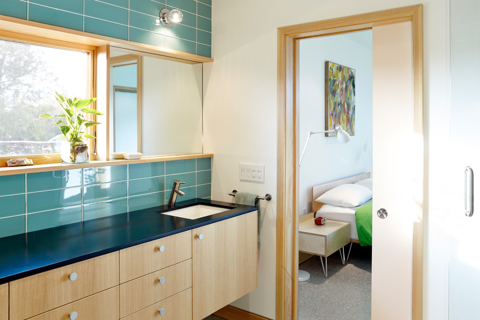 Modelo de cuarto de baño costero con lavabo bajoencimera, armarios con paneles lisos, puertas de armario de madera clara, baldosas y/o azulejos azules, paredes blancas y ventanas