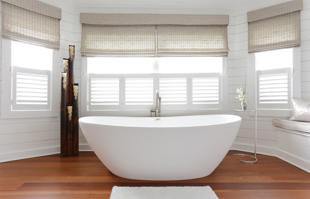 Foto de cuarto de baño marinero con bañera exenta, paredes blancas, suelo de madera en tonos medios y ventanas