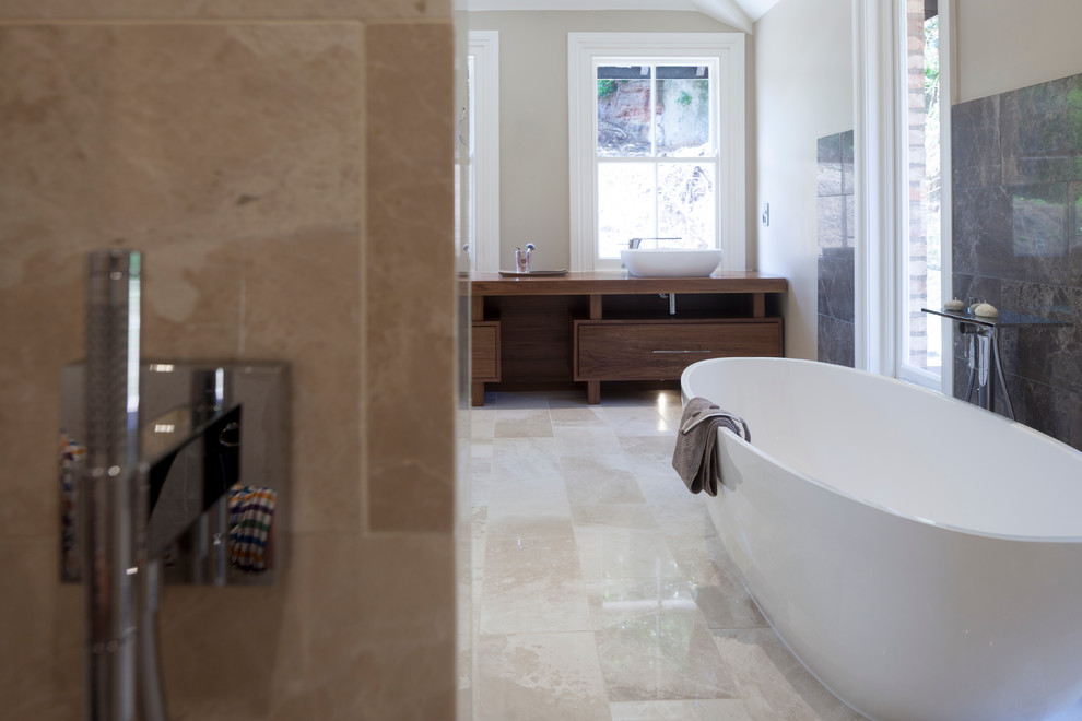 Photo of a large contemporary bathroom in Cheshire with a console sink, flat-panel cabinets, dark wood cabinets, wooden worktops, a freestanding bath, a walk-in shower, a wall mounted toilet, grey tiles, beige walls and limestone flooring.