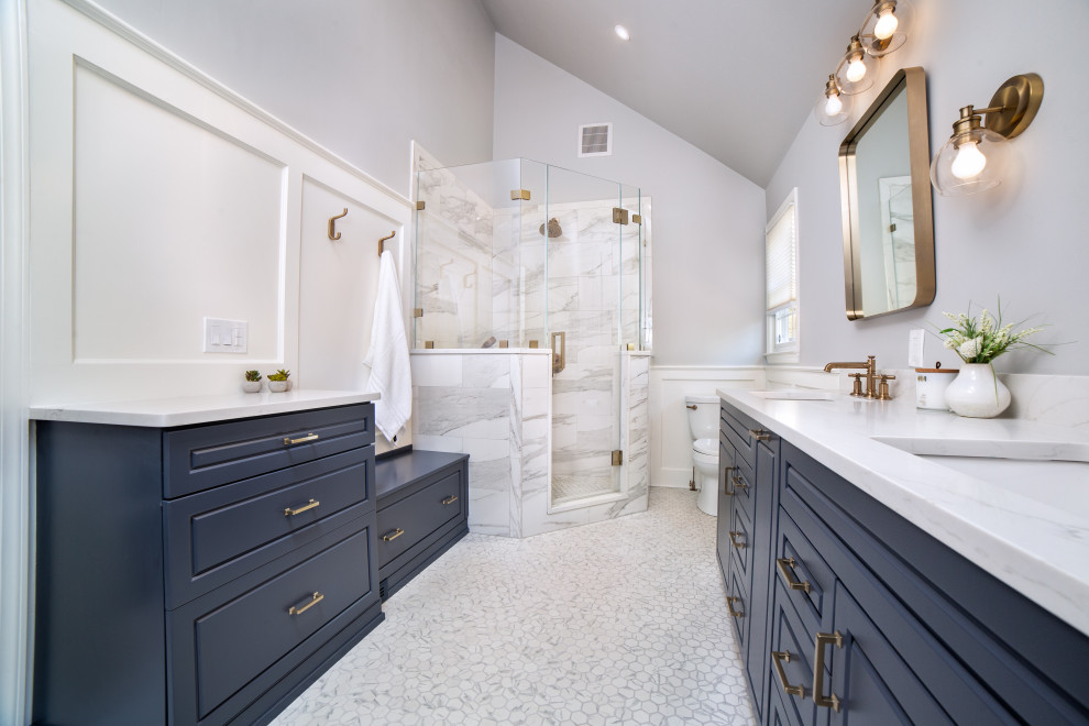 Пример оригинального дизайна: большая главная ванная комната в стиле неоклассика (современная классика) с фасадами с выступающей филенкой, синими фасадами, угловым душем, раздельным унитазом, белой плиткой, керамической плиткой, серыми стенами, полом из керамической плитки, врезной раковиной, столешницей из искусственного кварца, белым полом, душем с распашными дверями, белой столешницей, сиденьем для душа, тумбой под две раковины, встроенной тумбой, сводчатым потолком и панелями на стенах