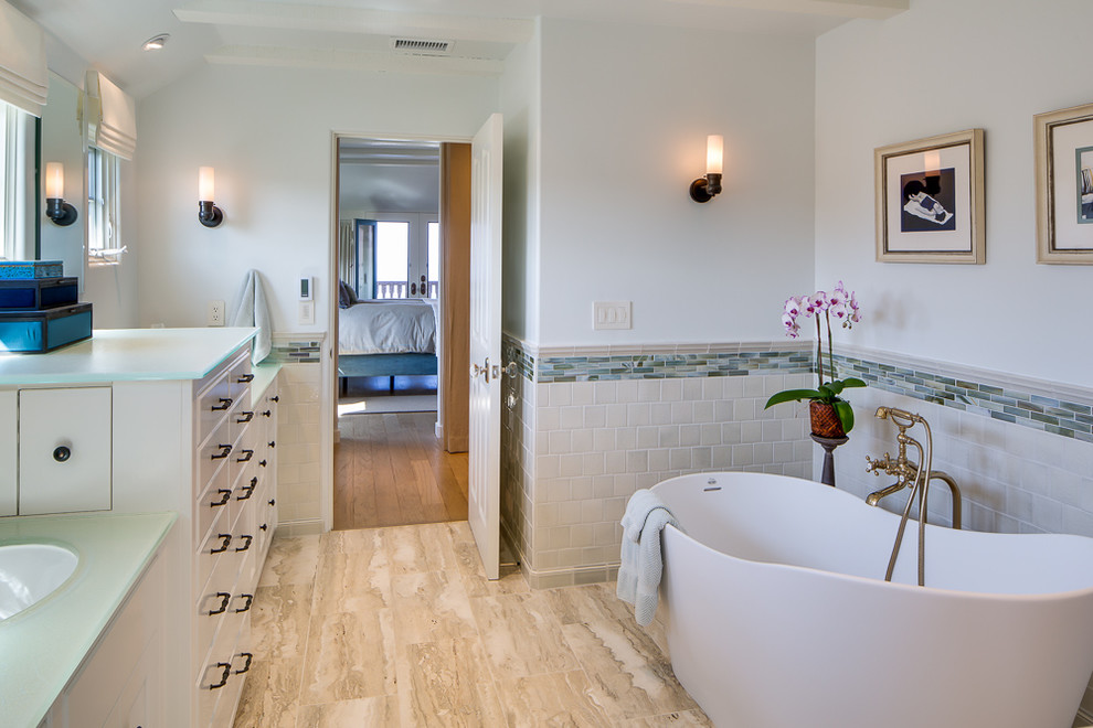 Идея дизайна: главная ванная комната в морском стиле с врезной раковиной, белыми фасадами, стеклянной столешницей, отдельно стоящей ванной, разноцветной плиткой, удлиненной плиткой, белыми стенами, полом из травертина и фасадами островного типа