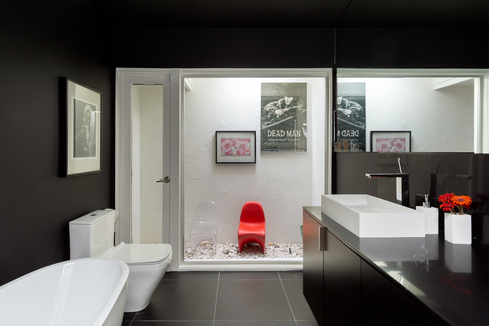 This is an example of a medium sized contemporary bathroom in Los Angeles with flat-panel cabinets, black cabinets, a freestanding bath, a one-piece toilet, black walls, ceramic flooring, a vessel sink, engineered stone worktops, black floors and black worktops.