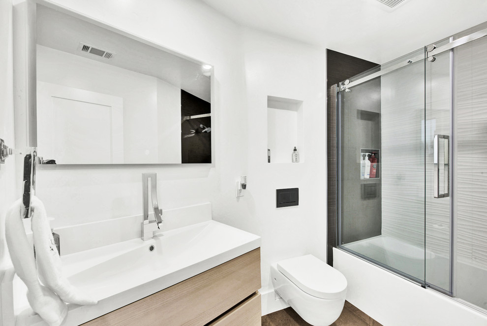 Inspiration for a medium sized contemporary shower room bathroom in San Francisco with flat-panel cabinets, light wood cabinets, an alcove bath, a shower/bath combination, a wall mounted toilet, white tiles, ceramic tiles, white walls, dark hardwood flooring, an integrated sink, solid surface worktops, brown floors, a sliding door and white worktops.