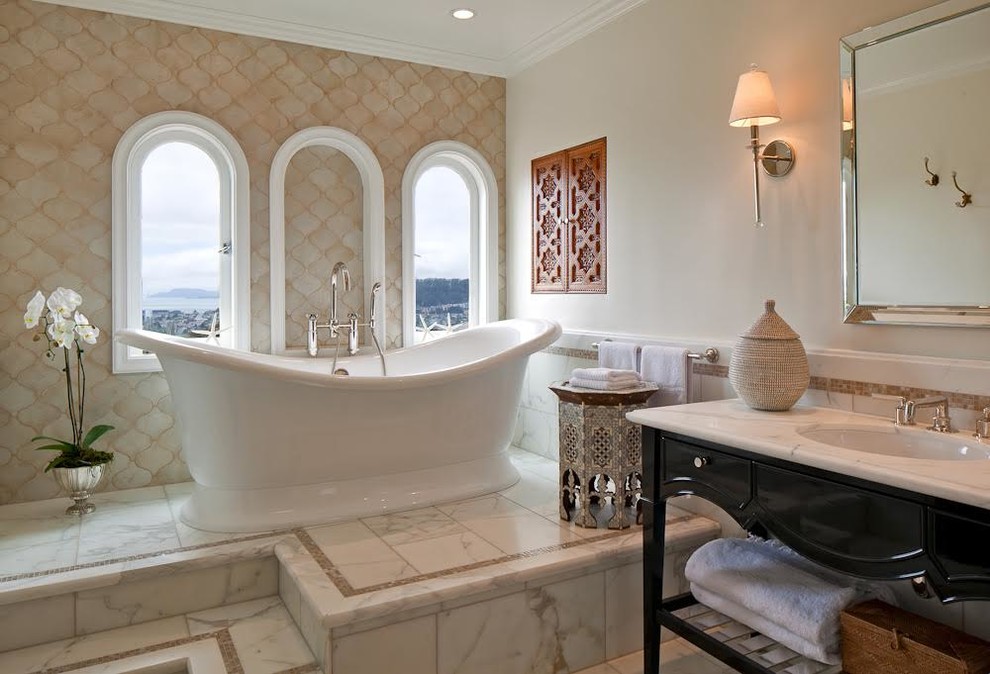 Medelhavsstil inredning av ett en-suite badrum, med svarta skåp, ett fristående badkar, beige väggar, ett undermonterad handfat, beige kakel, glaskakel, marmorbänkskiva och luckor med infälld panel