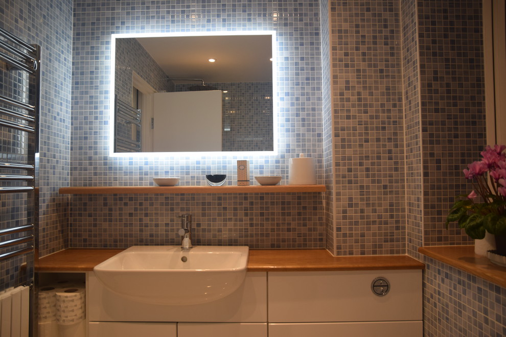 Small contemporary family bathroom in London with a built-in bath, a shower/bath combination, a wall mounted toilet, blue tiles, mosaic tiles, blue walls, porcelain flooring, a submerged sink and wooden worktops.