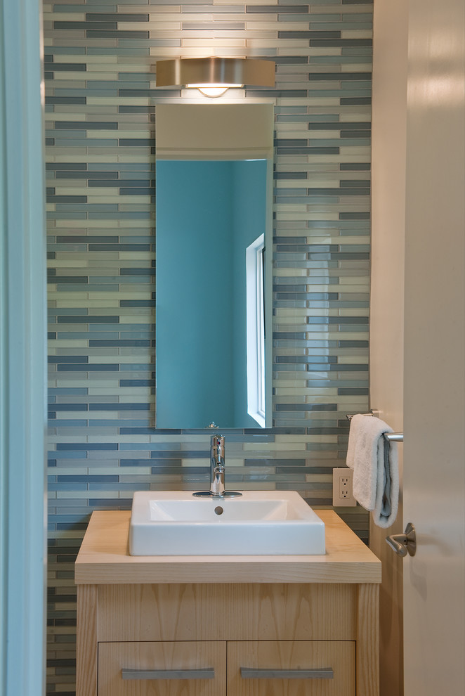 Modelo de cuarto de baño actual con lavabo sobreencimera, armarios con paneles lisos, puertas de armario de madera clara, encimera de madera, baldosas y/o azulejos azules y baldosas y/o azulejos de vidrio