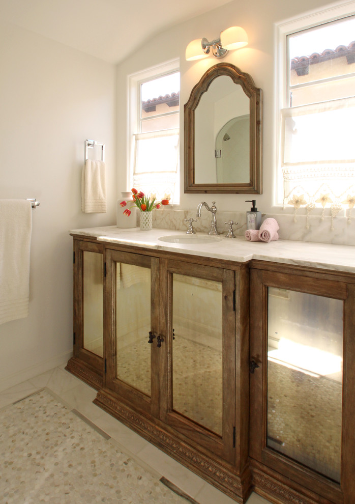 Modelo de cuarto de baño tradicional renovado con lavabo bajoencimera, armarios tipo vitrina, puertas de armario de madera oscura, encimera de mármol, paredes blancas y suelo de mármol