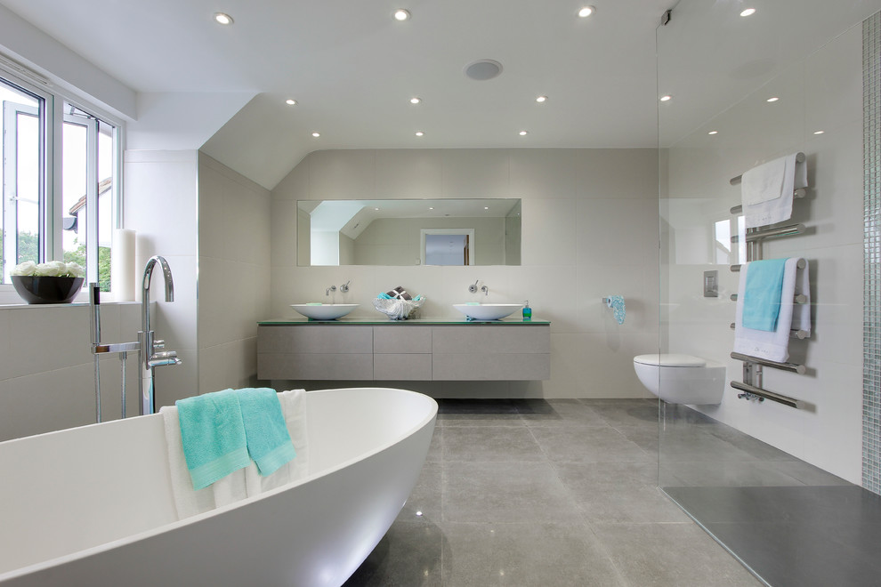 Modern inredning av ett en-suite badrum, med ett fristående handfat, släta luckor, grå skåp, ett fristående badkar, en kantlös dusch, en vägghängd toalettstol, grå kakel, porslinskakel, vita väggar, klinkergolv i porslin och bänkskiva i glas