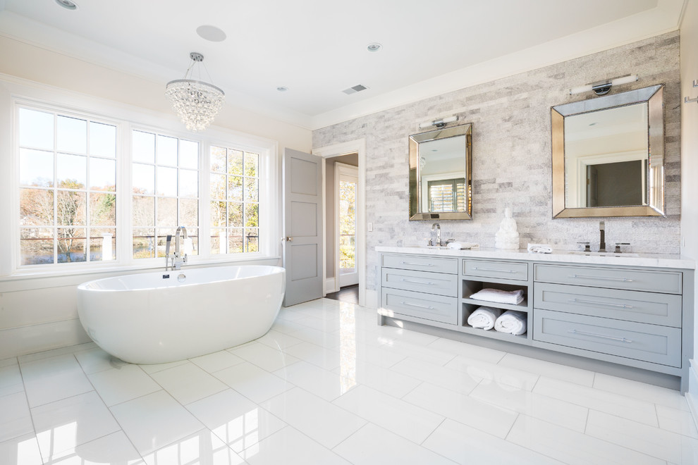 Cette photo montre une salle de bain principale chic avec un placard à porte shaker, des portes de placard grises, une baignoire indépendante, un carrelage gris, un mur beige, un lavabo encastré et un sol blanc.