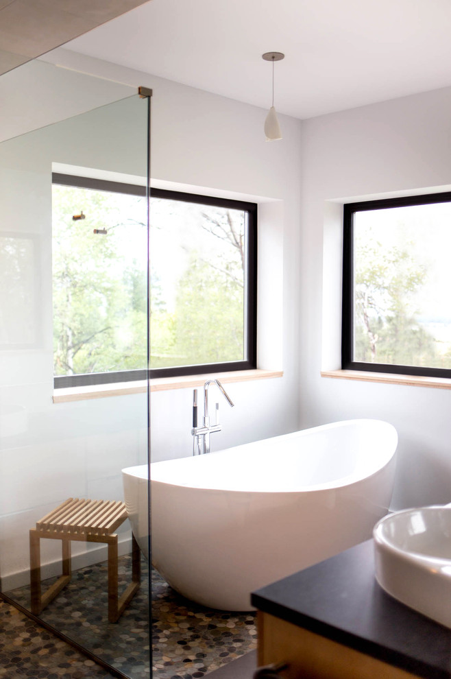 На фото: главная ванная комната среднего размера в стиле модернизм с настольной раковиной, плоскими фасадами, светлыми деревянными фасадами, отдельно стоящей ванной, угловым душем, инсталляцией, серой плиткой, белыми стенами и полом из галечной плитки