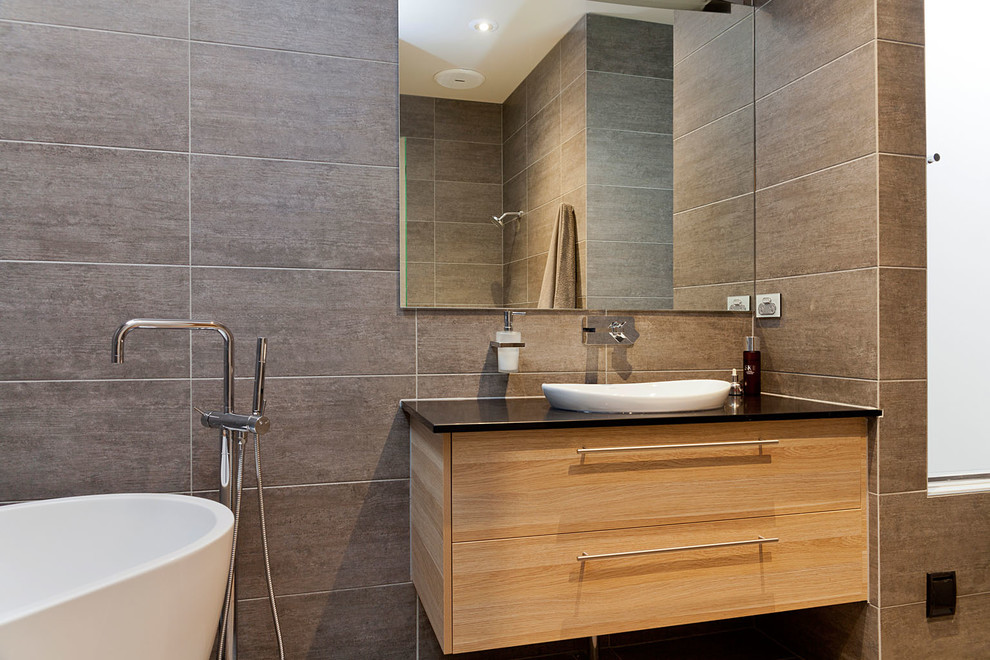 Modelo de cuarto de baño contemporáneo con armarios con paneles con relieve, puertas de armario de madera clara, encimera de granito, bañera exenta, ducha abierta, sanitario de pared y baldosas y/o azulejos grises
