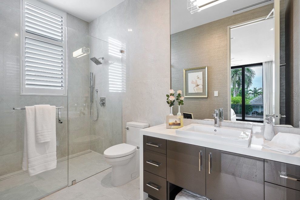 Photo of a large modern ensuite bathroom in Miami with flat-panel cabinets, dark wood cabinets, a freestanding bath, white tiles, stone slabs, beige walls, porcelain flooring, a built-in sink, onyx worktops, white floors, a hinged door, white worktops, a built-in shower and a one-piece toilet.
