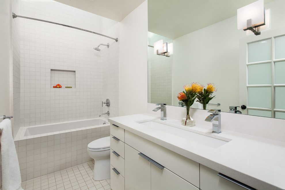 Bild på ett litet funkis badrum för barn, med ett avlångt handfat, ett badkar i en alkov, en dusch/badkar-kombination, vit kakel, släta luckor, vita skåp, en toalettstol med separat cisternkåpa, keramikplattor, vita väggar, klinkergolv i keramik, bänkskiva i kvarts, vitt golv och dusch med duschdraperi