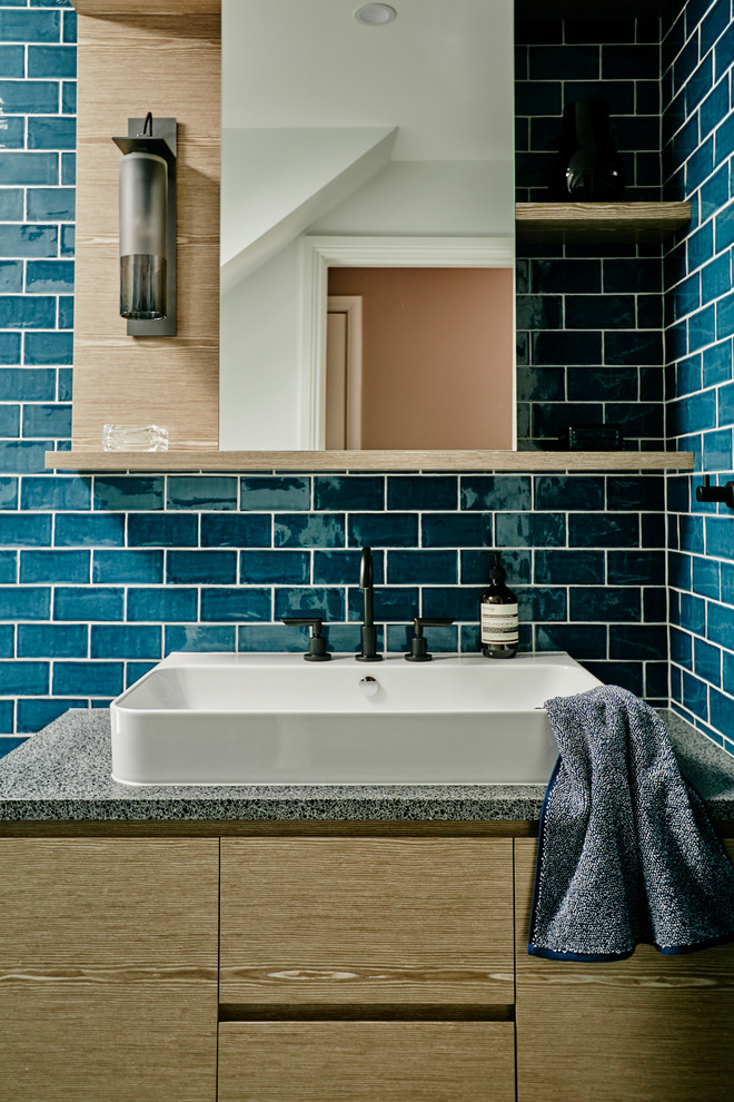 Bild på ett litet funkis badrum, med skåp i mellenmörkt trä, en dusch i en alkov, en vägghängd toalettstol, blå kakel, keramikplattor, blå väggar, cementgolv, ett avlångt handfat, bänkskiva i terrazo, svart golv och med dusch som är öppen