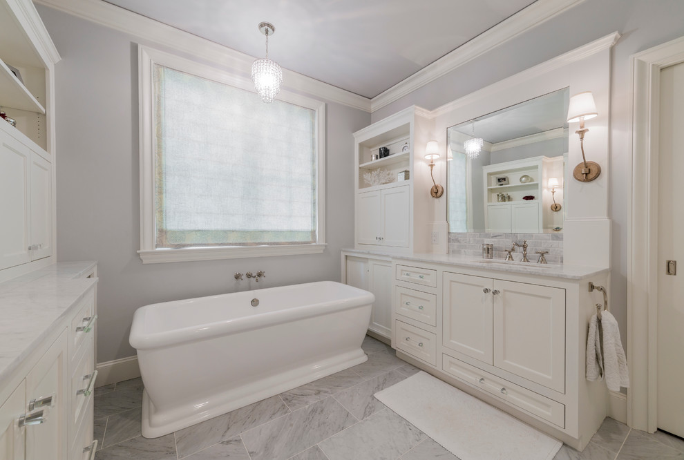 Cette image montre une grande salle de bain principale traditionnelle avec un placard à porte shaker, des portes de placard blanches, une baignoire indépendante, un mur gris, un sol en carrelage de porcelaine, un lavabo encastré et un plan de toilette en marbre.