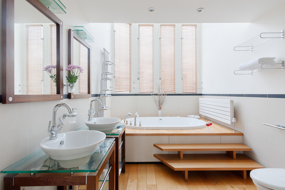 ロンドンにある中くらいなコンテンポラリースタイルのおしゃれな浴室 (中間色木目調キャビネット、アンダーマウント型浴槽、シャワー付き浴槽	、一体型トイレ	、磁器タイル、白い壁、淡色無垢フローリング、横長型シンク、ガラスの洗面台) の写真