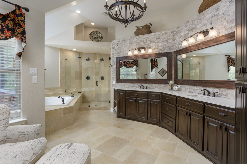 Large mediterranean ensuite bathroom in Dallas with raised-panel cabinets, dark wood cabinets, a built-in bath, a corner shower, a one-piece toilet, multi-coloured tiles, travertine tiles, beige walls, travertine flooring, a submerged sink, marble worktops, beige floors, a hinged door and white worktops.