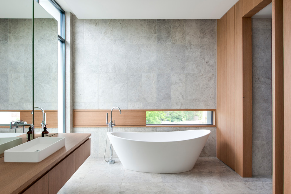 Foto på ett funkis brun badrum, med släta luckor, skåp i mellenmörkt trä, ett fristående badkar, grå väggar, ett fristående handfat, träbänkskiva och grått golv