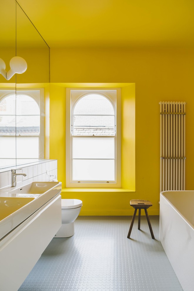 Modernes Badezimmer mit flächenbündigen Schrankfronten, weißen Schränken, freistehender Badewanne, Toilette mit Aufsatzspülkasten, weißen Fliesen, gelber Wandfarbe, Waschtischkonsole und grauem Boden in Sonstige