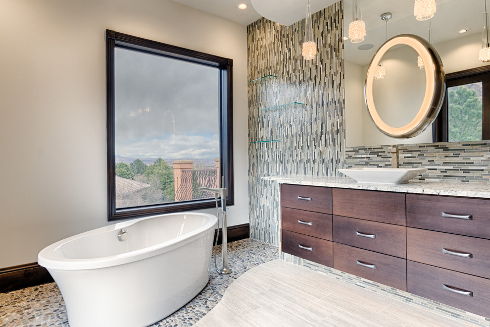 Photo of an expansive modern ensuite bathroom in Denver with flat-panel cabinets, dark wood cabinets, a freestanding bath, a two-piece toilet, multi-coloured tiles, glass sheet walls, beige walls, porcelain flooring, a vessel sink, granite worktops, beige floors and a hinged door.