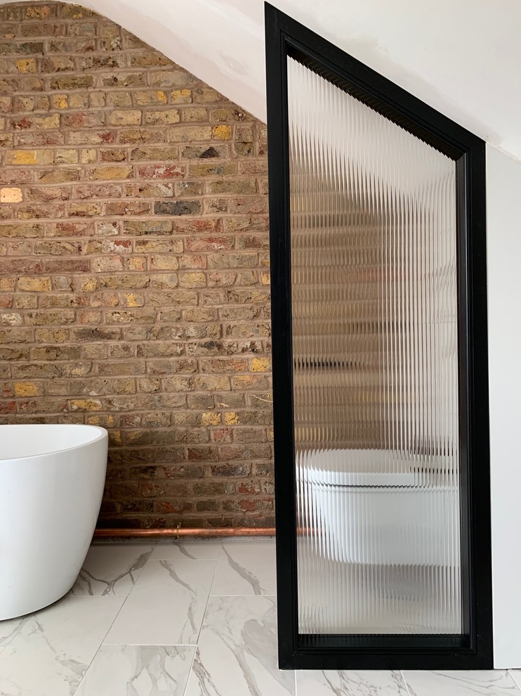 ロンドンにあるラグジュアリーな小さなインダストリアルスタイルのおしゃれなマスターバスルーム (置き型浴槽、一体型トイレ	、白いタイル、磁器タイル、茶色い壁、磁器タイルの床、白い床、三角天井、レンガ壁) の写真