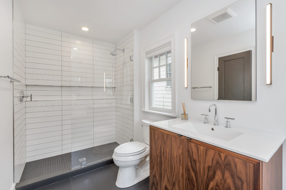 Bild på ett mellanstort industriellt badrum med dusch, med en toalettstol med separat cisternkåpa, svart kakel, vit kakel, klinkergolv i keramik, ett integrerad handfat, släta luckor, skåp i mellenmörkt trä, en dusch i en alkov, tunnelbanekakel, vita väggar, bänkskiva i akrylsten, ett fristående badkar, grått golv och med dusch som är öppen