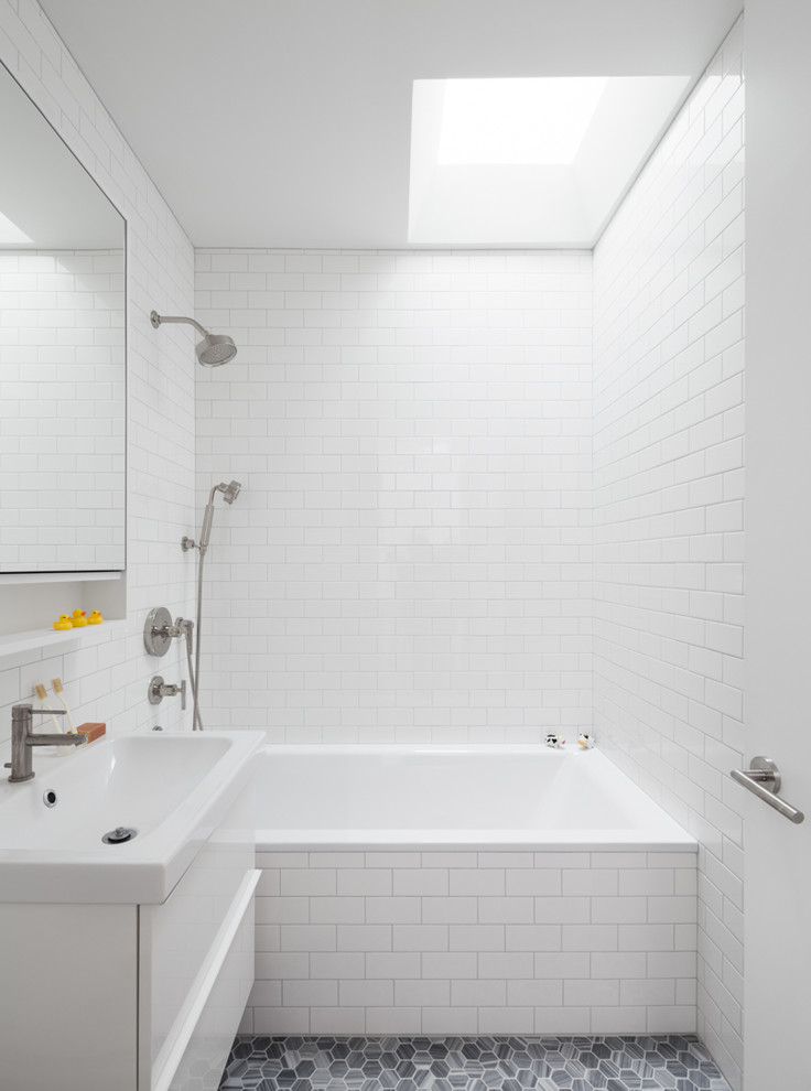 Modern inredning av ett litet badrum för barn, med släta luckor, vita skåp, ett platsbyggt badkar, en dusch/badkar-kombination, en vägghängd toalettstol, vit kakel, tunnelbanekakel, vita väggar, marmorgolv, ett väggmonterat handfat, bänkskiva i akrylsten, grått golv och med dusch som är öppen