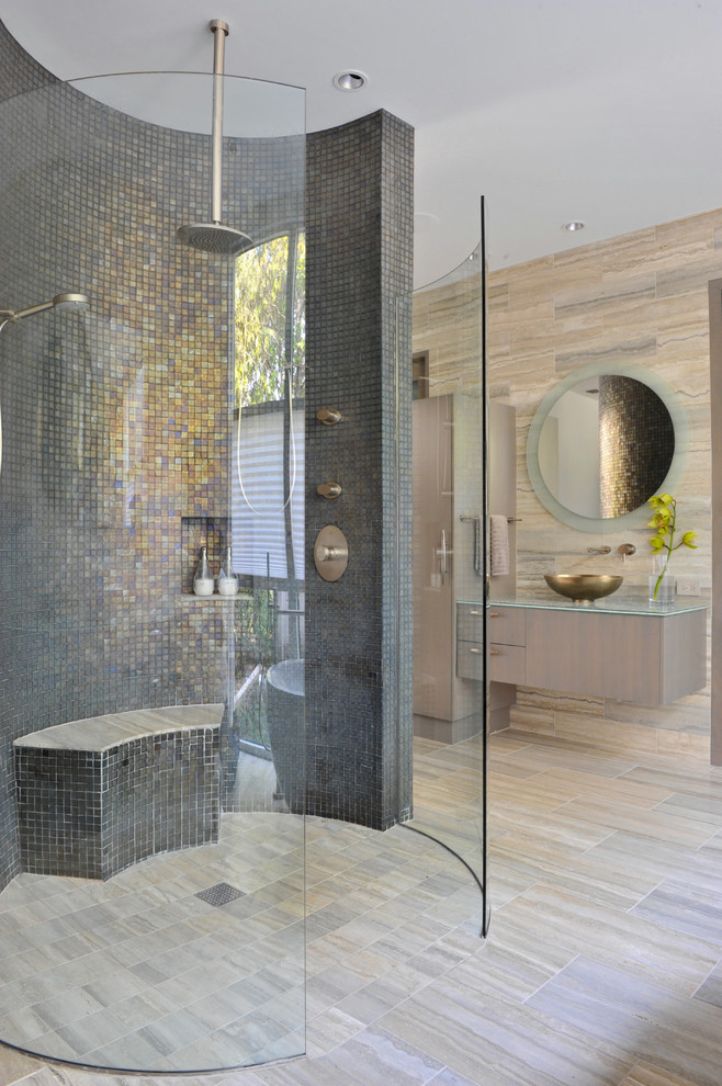 Cette image montre une très grande salle de bain design en bois clair avec une vasque, un placard à porte plane, un plan de toilette en verre, une baignoire indépendante, une douche à l'italienne, un carrelage de pierre, un carrelage gris, un mur gris et un sol en travertin.