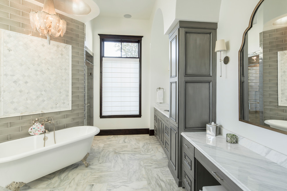 Aménagement d'une grande salle de bain principale victorienne avec un placard à porte affleurante, des portes de placard grises, une baignoire sur pieds, un carrelage gris, un carrelage en pâte de verre, un mur beige, un sol en marbre, un lavabo encastré, un plan de toilette en marbre et un sol blanc.