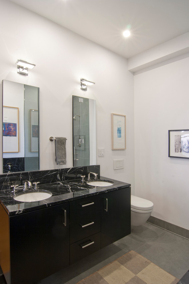 Aménagement d'une salle de bain principale moderne de taille moyenne avec un placard à porte plane, des portes de placard noires, une baignoire encastrée, une douche d'angle, WC suspendus, un carrelage noir, du carrelage en marbre, un mur blanc, un sol en carrelage de porcelaine, un lavabo encastré, un plan de toilette en marbre, un sol gris, une cabine de douche à porte battante et un plan de toilette noir.