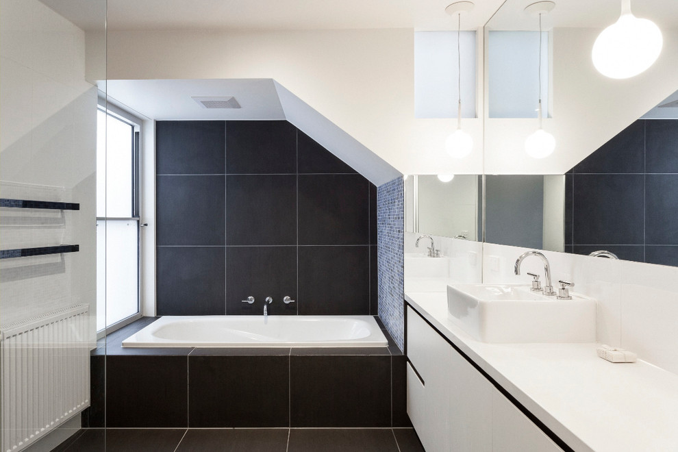 Modern inredning av ett badrum, med ett fristående handfat, släta luckor, vita skåp, ett platsbyggt badkar, svart kakel och vita väggar