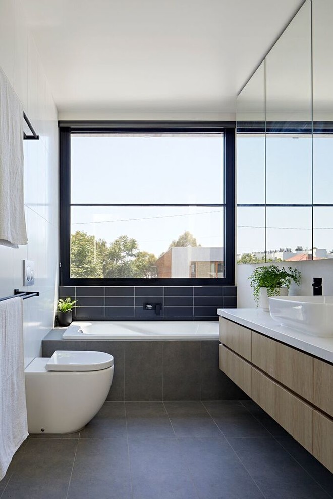 Bild på ett funkis vit vitt badrum, med släta luckor, skåp i ljust trä, ett platsbyggt badkar, en vägghängd toalettstol, svart kakel, grå kakel, vit kakel, vita väggar, ett fristående handfat och grått golv