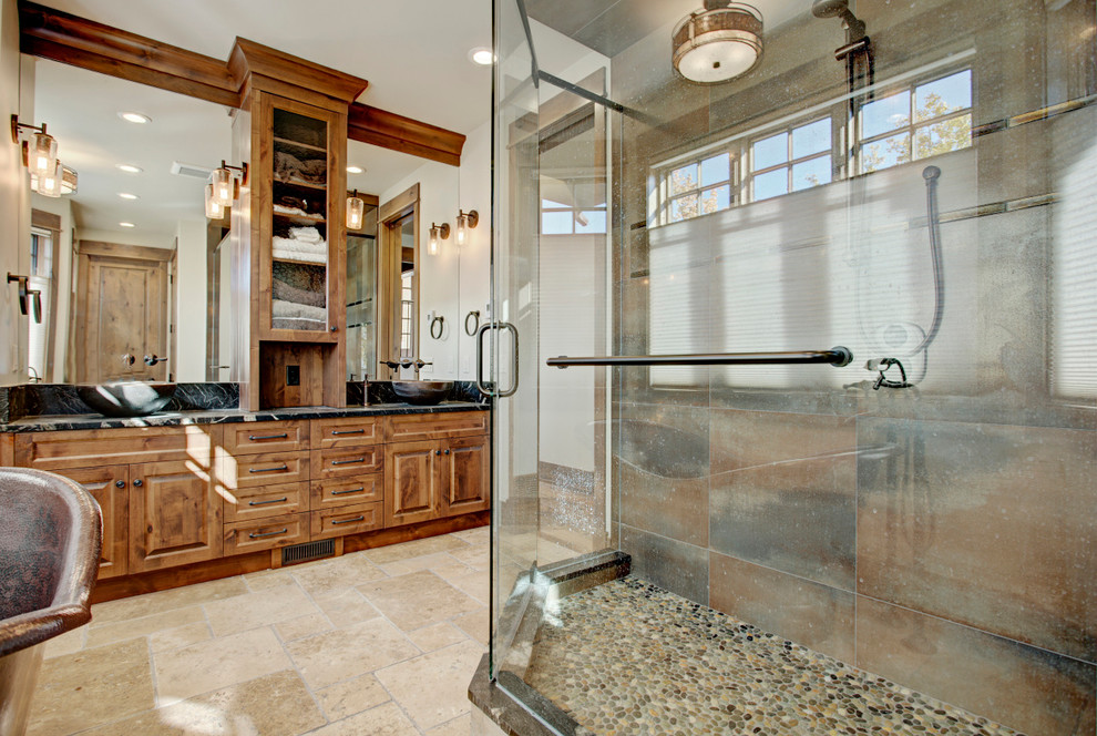 Design ideas for a large rustic ensuite bathroom in Calgary with raised-panel cabinets, medium wood cabinets, a freestanding bath, a built-in shower, a one-piece toilet, beige tiles, brown tiles, grey tiles, stone tiles, beige walls, travertine flooring, a submerged sink, granite worktops, beige floors and a hinged door.