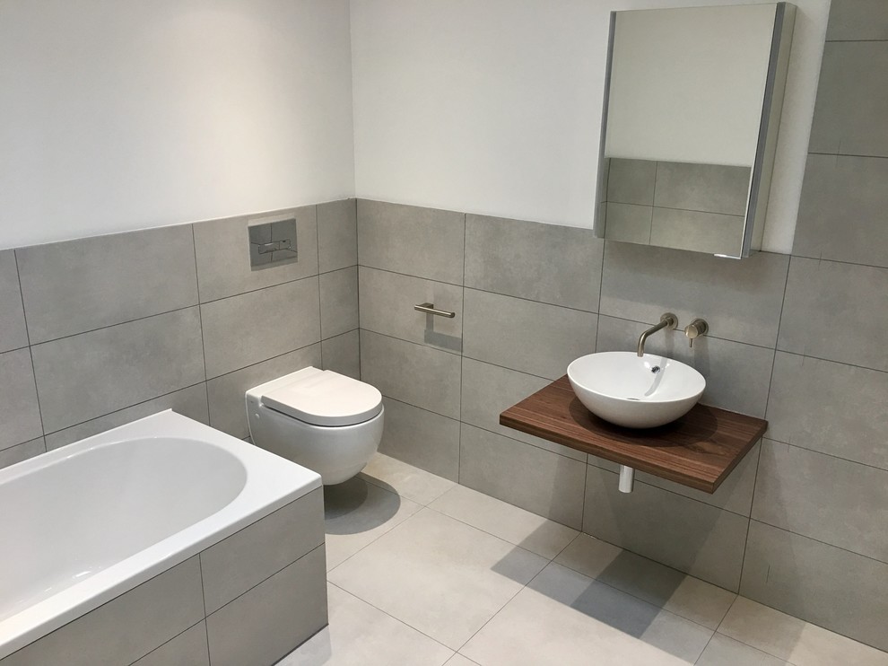 Medium sized contemporary ensuite wet room bathroom in Kent with flat-panel cabinets, a built-in bath, a wall mounted toilet, grey tiles, porcelain tiles, grey walls, porcelain flooring, a vessel sink, wooden worktops and grey floors.