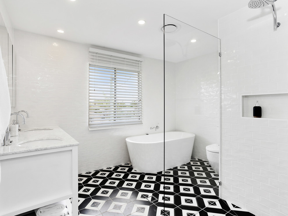 Foto på ett maritimt vit badrum, med vita skåp, ett fristående badkar, vit kakel, keramikplattor, vita väggar, ett nedsänkt handfat, marmorbänkskiva, flerfärgat golv och dusch med gångjärnsdörr
