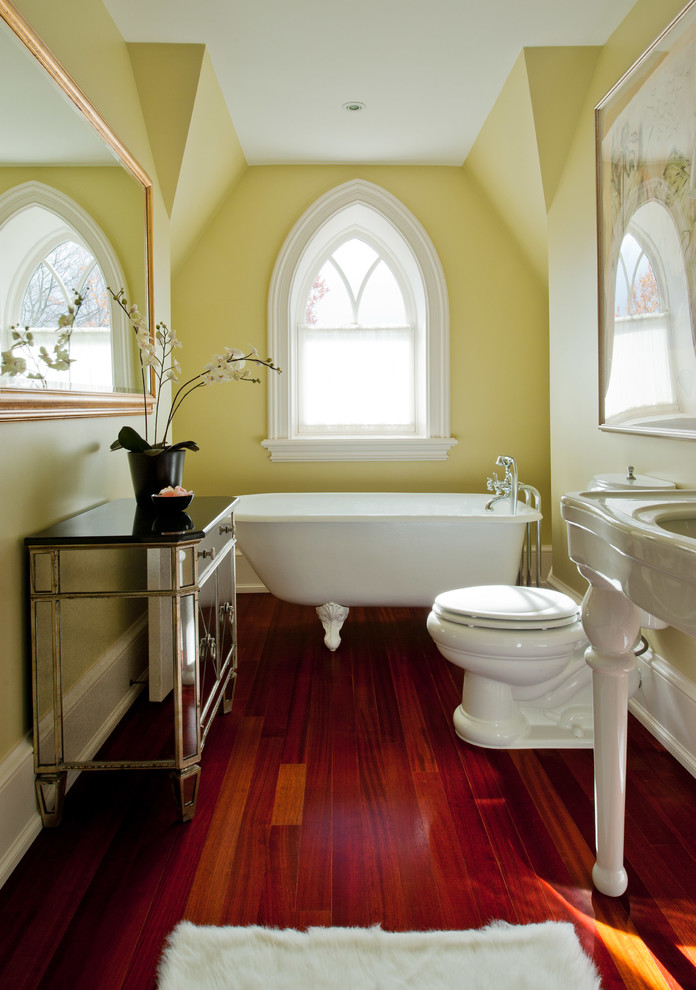 Idee per una stanza da bagno classica con lavabo a consolle, vasca con piedi a zampa di leone, pareti gialle, pavimento in legno massello medio e pavimento marrone