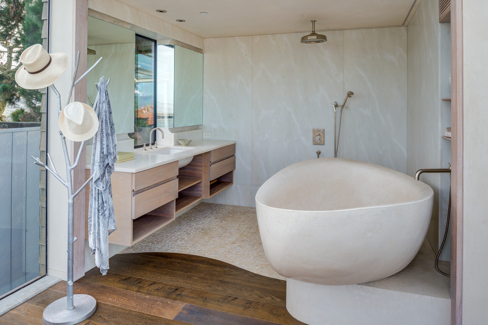 Cette image montre une salle de bain design en bois clair avec un placard à porte plane, une baignoire indépendante, une douche à l'italienne, un carrelage beige, un mur beige, un sol en galet, un lavabo encastré, un sol beige, aucune cabine et un plan de toilette blanc.
