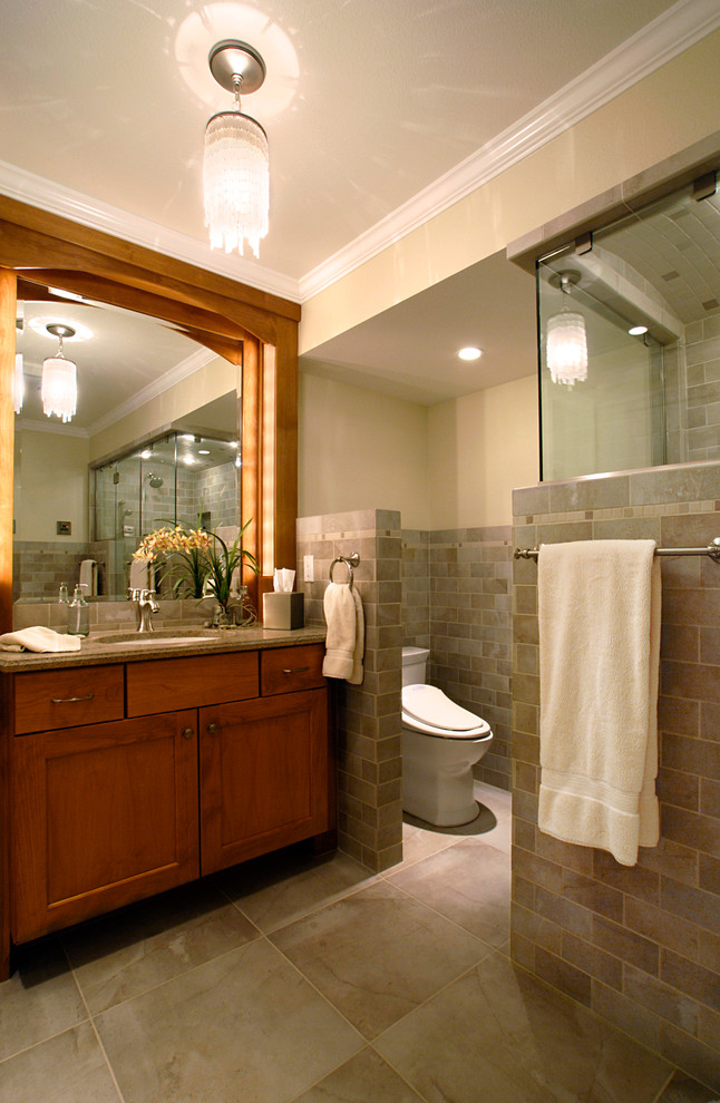 Cette image montre une salle de bain traditionnelle en bois brun de taille moyenne avec un placard à porte shaker, WC à poser, un carrelage gris, un carrelage de pierre, un mur beige, un sol en carrelage de porcelaine, un lavabo encastré et un plan de toilette en granite.