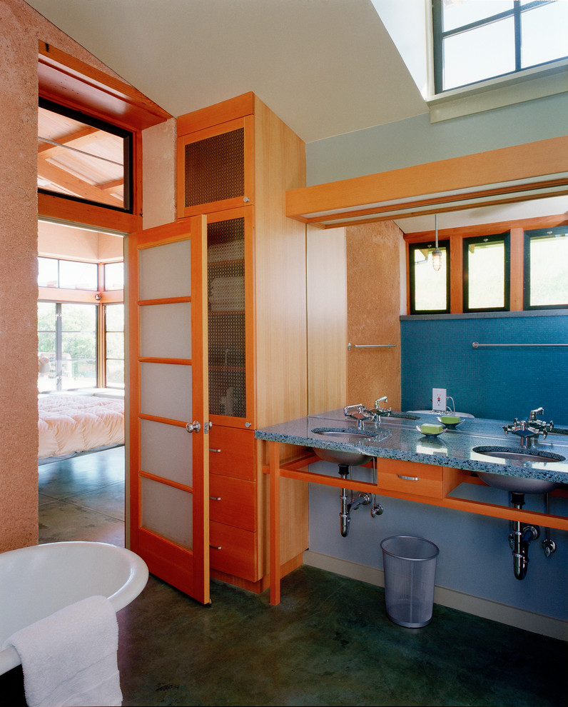 На фото: ванная комната в стиле модернизм с плоскими фасадами, светлыми деревянными фасадами, ванной на ножках, синими стенами, бетонным полом, врезной раковиной и столешницей из переработанного стекла
