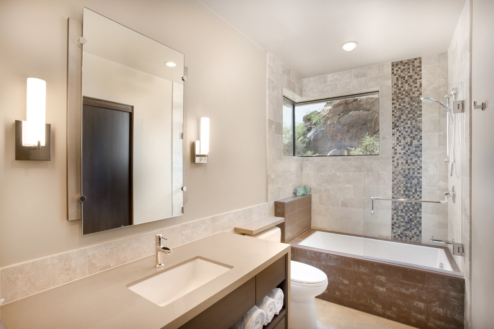 Exempel på ett stort modernt badrum, med släta luckor, skåp i mellenmörkt trä, ett badkar i en alkov, en dusch/badkar-kombination, en toalettstol med hel cisternkåpa, beige kakel, stenkakel, beige väggar, kalkstensgolv, ett undermonterad handfat och granitbänkskiva