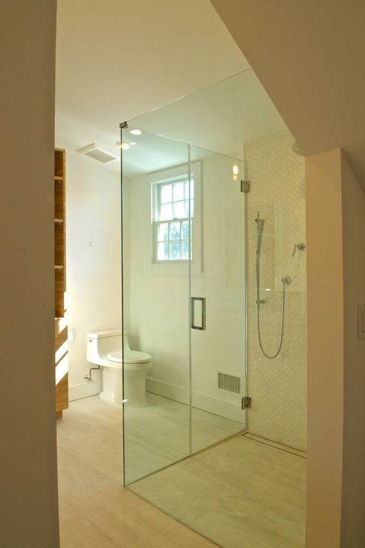 This is an example of a medium sized coastal ensuite bathroom in New York with flat-panel cabinets, light wood cabinets, a built-in shower, a one-piece toilet, beige tiles, metro tiles, white walls, porcelain flooring, a submerged sink, engineered stone worktops, grey floors, a hinged door and white worktops.