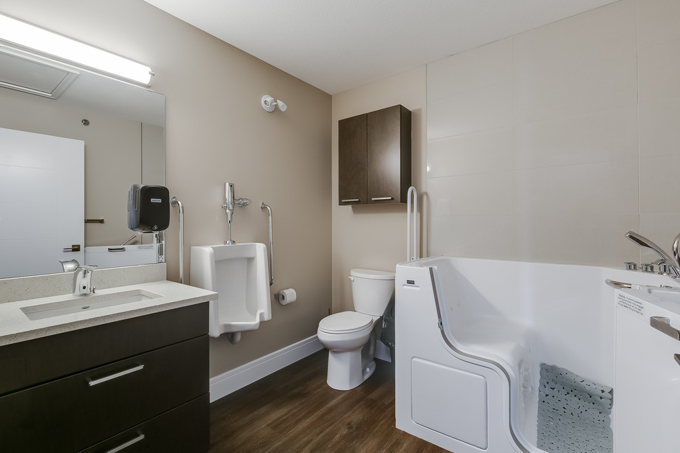 Bild på ett mellanstort funkis badrum, med släta luckor, skåp i mörkt trä, ett hörnbadkar, en dusch i en alkov, ett urinoar, vit kakel, beige väggar, vinylgolv, ett undermonterad handfat och bänkskiva i kvartsit