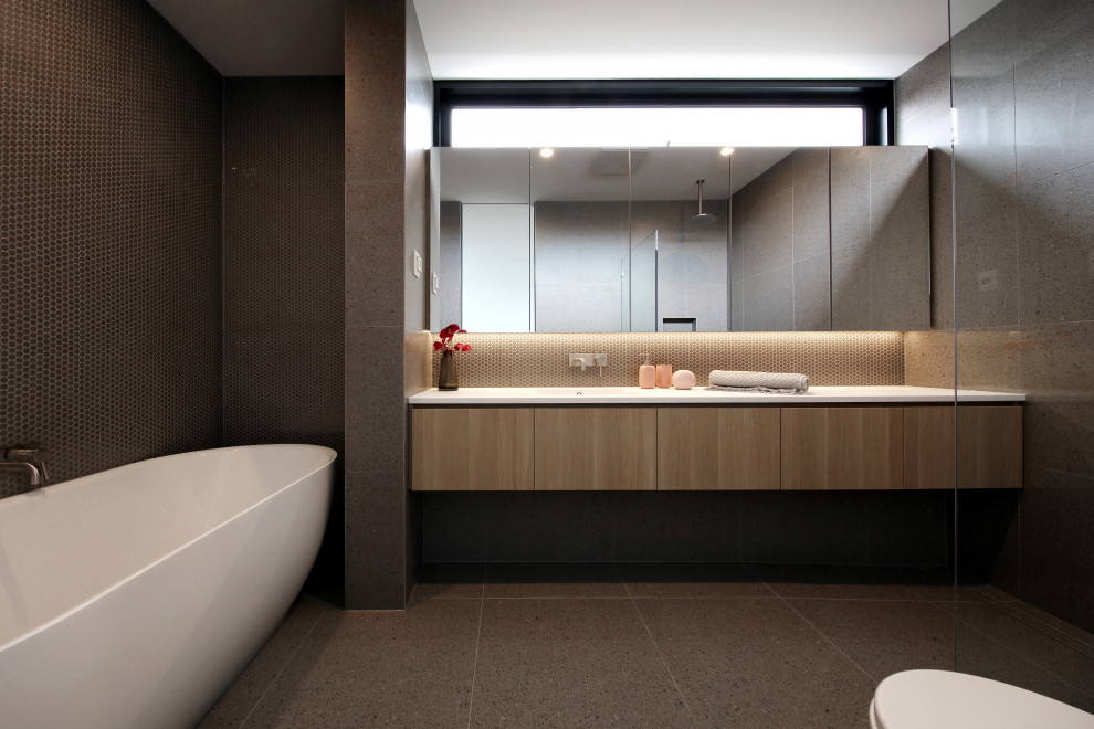 На фото: ванная комната в современном стиле с плоскими фасадами, фасадами цвета дерева среднего тона, отдельно стоящей ванной, серой плиткой, серым полом и белой столешницей