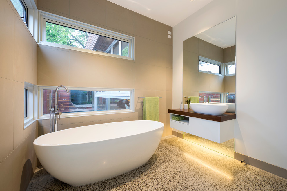 На фото: ванная комната в современном стиле с настольной раковиной, плоскими фасадами, белыми фасадами, отдельно стоящей ванной, бежевой плиткой, белыми стенами и бетонным полом с