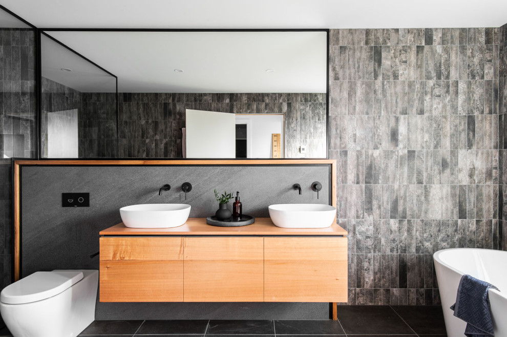 Cette photo montre une salle de bain principale tendance en bois brun avec un placard à porte plane, une baignoire indépendante, WC à poser, un carrelage gris, un mur gris, une vasque, un plan de toilette en bois, un sol gris, un plan de toilette marron, meuble double vasque et meuble-lavabo suspendu.