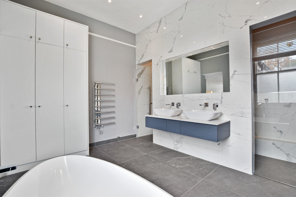 Design ideas for a large contemporary ensuite bathroom in London with flat-panel cabinets, blue cabinets, a freestanding bath, an alcove shower, grey tiles, white tiles, grey walls, a vessel sink, grey floors, a hinged door and white worktops.