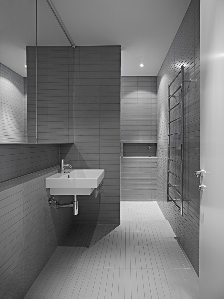 Modern inredning av ett badrum, med ett väggmonterat handfat, släta luckor, kaklad bänkskiva, en dusch i en alkov, grå kakel, stenhäll, grå väggar och klinkergolv i keramik